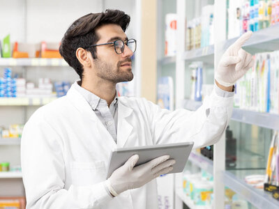 Eine Person trägt einen weißen Laborkittel und Handschuhe, hält ein Tablet in einer Hand und zeigt mit der anderen Hand auf Gegenstände in einem Regal. Der Hintergrund scheint Regale zu sein, die mit verschiedenen Produkten gefüllt sind, wahrscheinlich in einer Apotheke oder einem medizinischen Fachgeschäft.