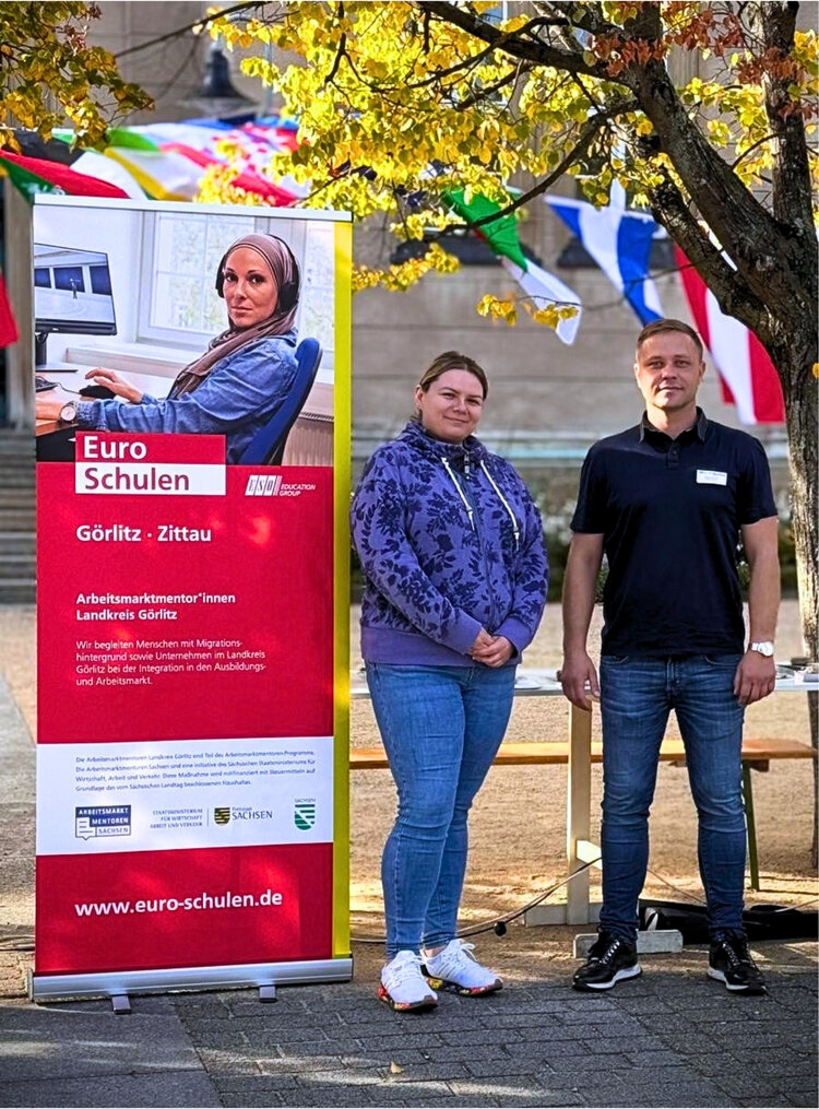 Zwei Mitarbeitende stehen bei einer Veranstaltung neben einem Roll-up der Euro-Schulen Görlitz-Zittau zum Projekt Arbeitsmarktmentor*innen im Landkreis Görlitz.