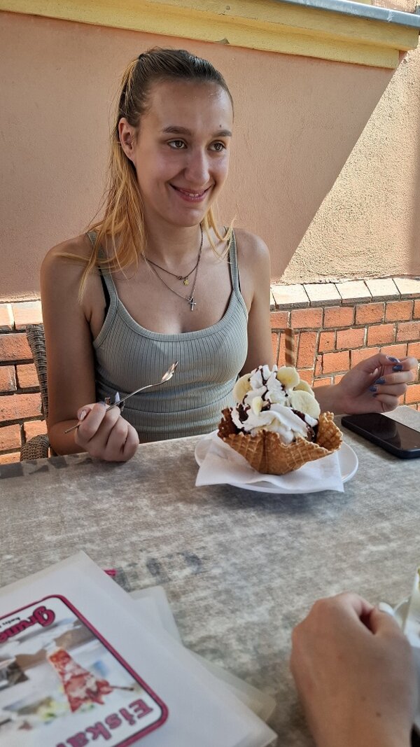 Eine junge Frau sitzt lächelnd vor einem großen Eisbecher in einer Waffelschale, garniert mit Sahne, Banane und Schokoladensoße.