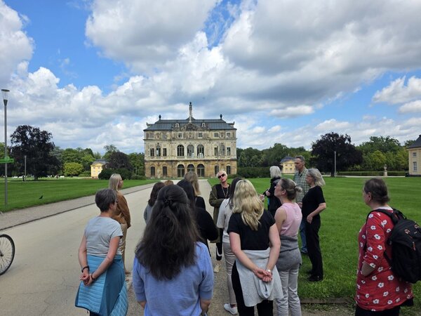 Eine Gruppe von Menschen steht auf dem Rasen und einem Weg in einem Park und nehmen an einer Stadtführung teil. Im Hintergrund sind Bäume und ein historisches Gebäude zu sehen.