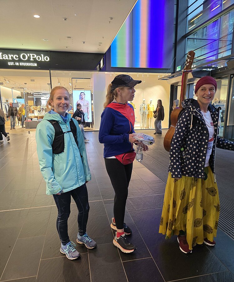 Euro-Schulen & Euro Akademie bei der Rewe Team Challenge 2025 Drei Frauen in Sportkleidung stehen in einem Einkaufszentrum vor dem Marc O’Polo Store. Eine trägt eine Gitarre, die anderen zwei tragen Sportjacken und Laufkleidung.