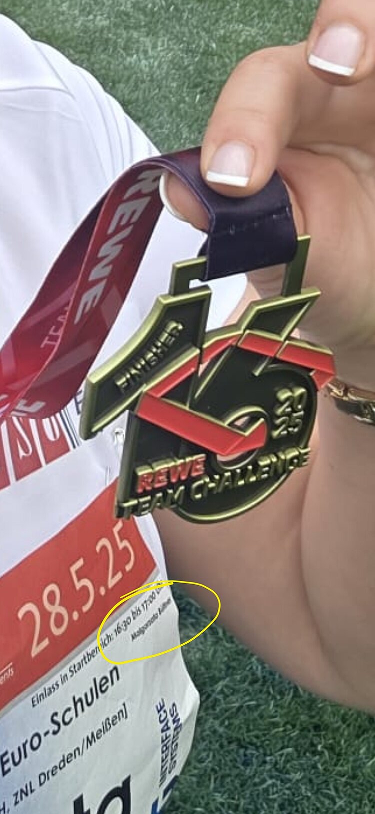 Euro-Schulen & Euro Akademie bei der Rewe Team Challenge 2025 Nahaufnahme einer Hand, die die REWE Team Challenge 2025 Finisher-Medaille hält. Im Hintergrund sieht man das Startnummernfeld mit Veranstaltungsinfos.