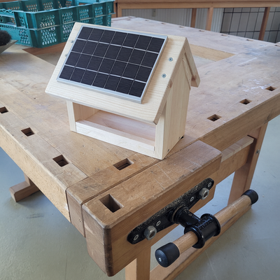 Ein kleines Häuschen aus hellem Holz steht auf einer Werkbank; auf dem schrägen Dach ist ein rechteckiges schwarzes Solarpanel montiert, das das Modell wie ein kleines solarbetriebenes Häuschen wirken lässt.