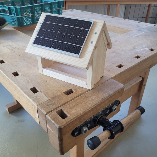 Ein kleines Häuschen aus hellem Holz steht auf einer Werkbank; auf dem schrägen Dach ist ein rechteckiges schwarzes Solarpanel montiert, das das Modell wie ein kleines solarbetriebenes Häuschen wirken lässt.