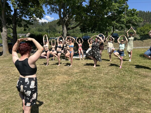 Auf einer Wiese praktiziert eine Gruppe junger Menschen Yoga: sie alle stehen auf einem Bein, halten das andere Bein angewinkelt und halten die Arme im Bogen über ihre Köpfe.