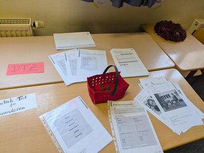 Auf einem hellen Holztisch liegen mehrere Stapel von ausgedruckten Arbeitsblättern und Formularen, teilweise in Klarsichthüllen. In der Mitte steht ein roter Kunststoffkorb mit Griff und einigen kleinen Gegenständen darin. Links auf dem Tisch liegt ein rosafarbenes Schild mit der Aufschrift 'DTZ', daneben ein weißes Blatt mit der Aufschrift 'Deutsch Test für Zuwanderer'. Im Hintergrund ist ein Heizkörper zu sehen, darüber hängt ein Vorhang. Rechts am Bildrand liegt ein dunkler Kranz oder eine Dekoration.