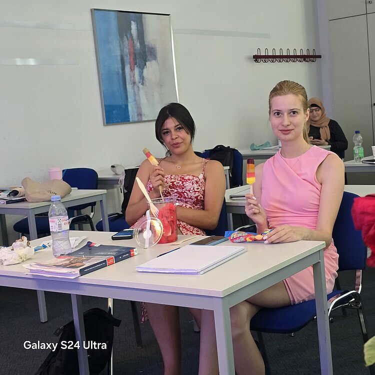 Heiße Köpfe, kühles Eis – Sprachkurse bei 35 Grad! Zwei junge Frauen sitzen in einem Klassenzimmer an einem Tisch und genießen ein Eis am Stiel. Auf dem Tisch liegen Hefte, Stifte und weitere Lernmaterialien. Im Hintergrund sind weitere Tische und ein abstraktes Gemälde an der Wand zu sehen.