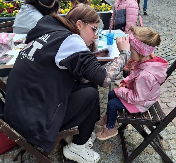 Eine junge Frau schminkt das Gesicht eines kleinen Mädchens, das auf einem Stuhl sitzt. Im Hintergrund sind Blumen und andere Kinder zu sehen.
