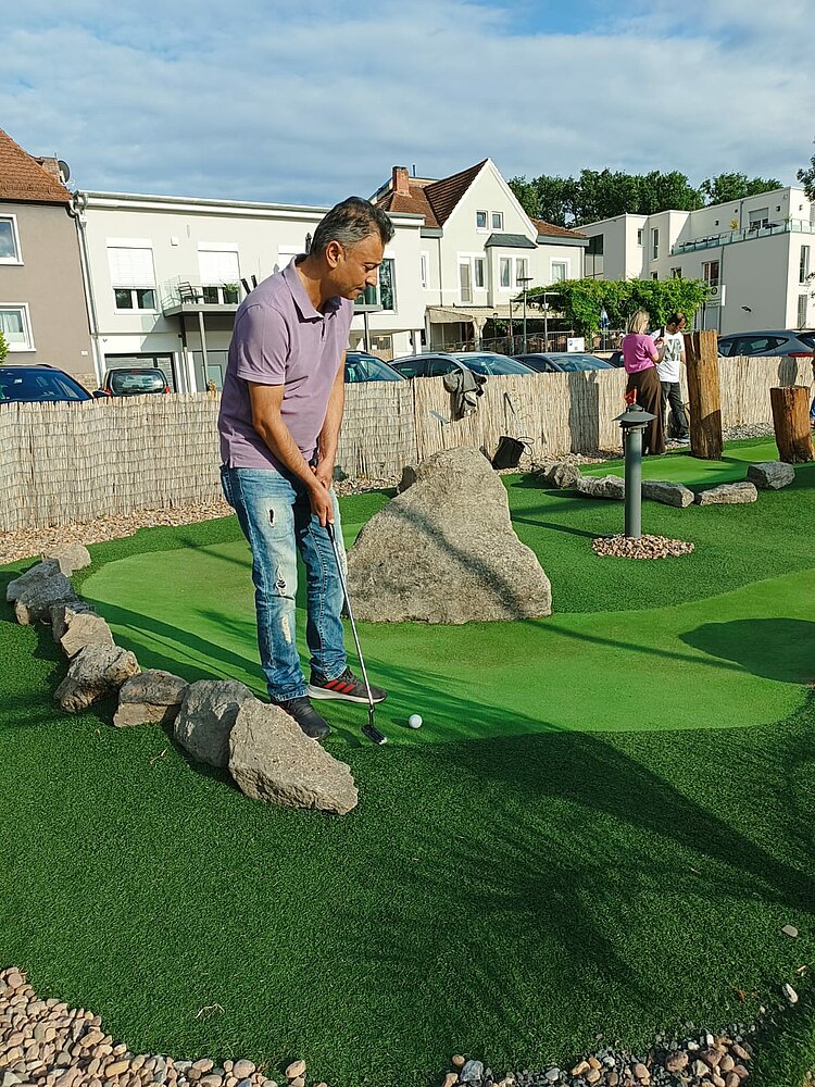 Ein Mann in einem lila Polohemd und Jeans steht auf einem Minigolfplatz. Er hält einen Putter und bereitet sich darauf vor, einen Golfball über eine künstliche Grünfläche mit Hindernissen aus großen Steinen zu spielen. Im Hintergrund sind weitere Spieler, ein Zaun aus Weidengeflecht und Wohnhäuser zu sehen.