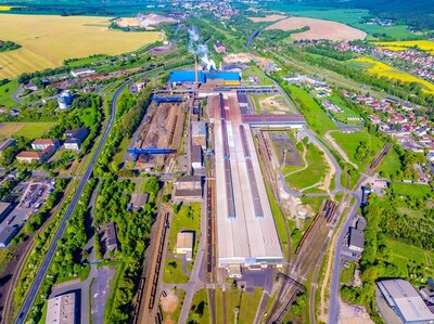 Eine große Industrieanlage des Stahlwerks in Thüringen mit mehreren langgestreckten Hallen und Schornsteinen liegt inmitten einer grünen Landschaft. Eisenbahngleise verlaufen parallel zu den Gebäuden und führen in verschiedene Richtungen. Im Hintergrund sind Felder, Wälder und eine Siedlung mit Wohnhäusern zu sehen. Rauch steigt aus einem der Schornsteine auf. Die Aufnahme zeigt das gesamte Gelände aus der Vogelperspektive.