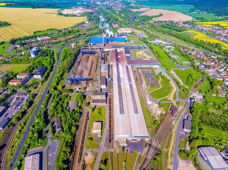 Eine große Industrieanlage des Stahlwerks in Thüringen mit mehreren langgestreckten Hallen und Schornsteinen liegt inmitten einer grünen Landschaft. Eisenbahngleise verlaufen parallel zu den Gebäuden und führen in verschiedene Richtungen. Im Hintergrund sind Felder, Wälder und eine Siedlung mit Wohnhäusern zu sehen. Rauch steigt aus einem der Schornsteine auf. Die Aufnahme zeigt das gesamte Gelände aus der Vogelperspektive.