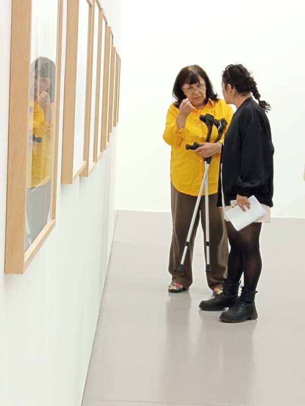 Dozentin im gelben Hemd erklärt einer Teilnehmerin mit Unterlagen und Gehstützen ein gerahmtes Kunstwerk an der weißen Wand; weitere Bilder hängen in einer Reihe daneben.