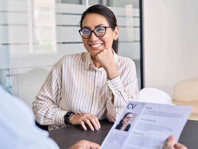 Eine junge Frau mit Brille und einem gestreiften, hellen Hemd sitzt lächelnd an einem Tisch in einem Büro oder Besprechungsraum. Sie wirkt selbstbewusst und entspannt, während sie ihr Kinn auf die Hand stützt. Vor ihr liegt ein Lebenslauf (CV) mit ihrem Foto und persönlichen Informationen, den eine andere Person, deren Arm zu sehen ist, in der Hand hält. Der Raum wirkt modern und hell, mit großen Fenstern im Hintergrund. Die Szene vermittelt den Eindruck eines Vorstellungsgesprächs oder einer beruflichen Besprechung.