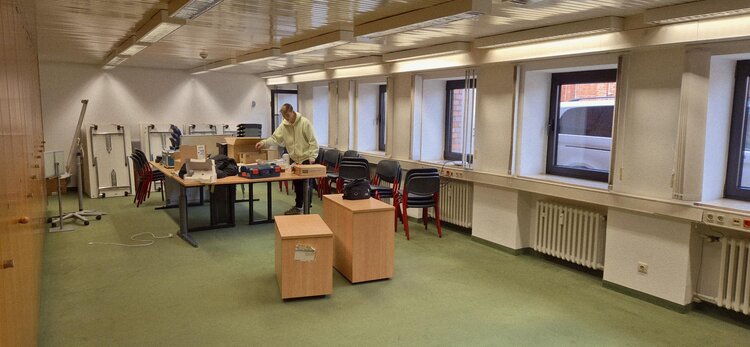 Großer Raum in Umzugsvorbereitung: gestapelte Stühle und Tische, Kartons und Material auf einem Tisch; eine Person packt und sortiert.