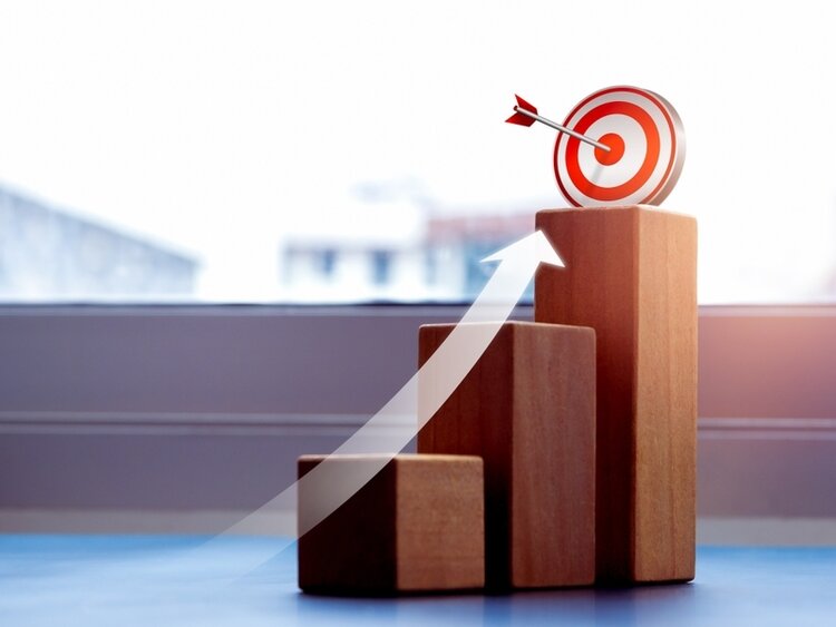 Drei Holzblöcke repräsentieren die Stufen, ein darübergelegter weißer Pfeil symbolisiert den Wachstum. Auf dem obersten Block steht eine weiß-rote Zielschiebe, in deren Mitte ein Dartpfeil steckt.