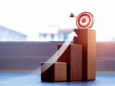 Drei Holzblöcke repräsentieren die Stufen, ein darübergelegter weißer Pfeil symbolisiert den Wachstum. Auf dem obersten Block steht eine weiß-rote Zielschiebe, in deren Mitte ein Dartpfeil steckt.