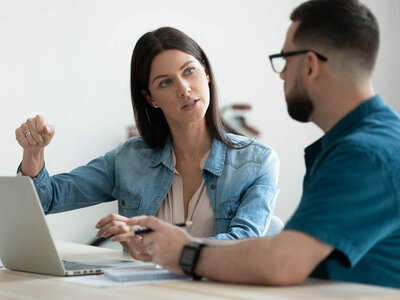 Zwei Personen sitzen an einem Tisch und führen eine Diskussion. Eine Person trägt eine Jeansjacke und gestikuliert mit der Hand, während die andere Person eine Brille trägt und einen Stift hält. Vor ihnen auf dem Tisch steht ein offener Laptop.