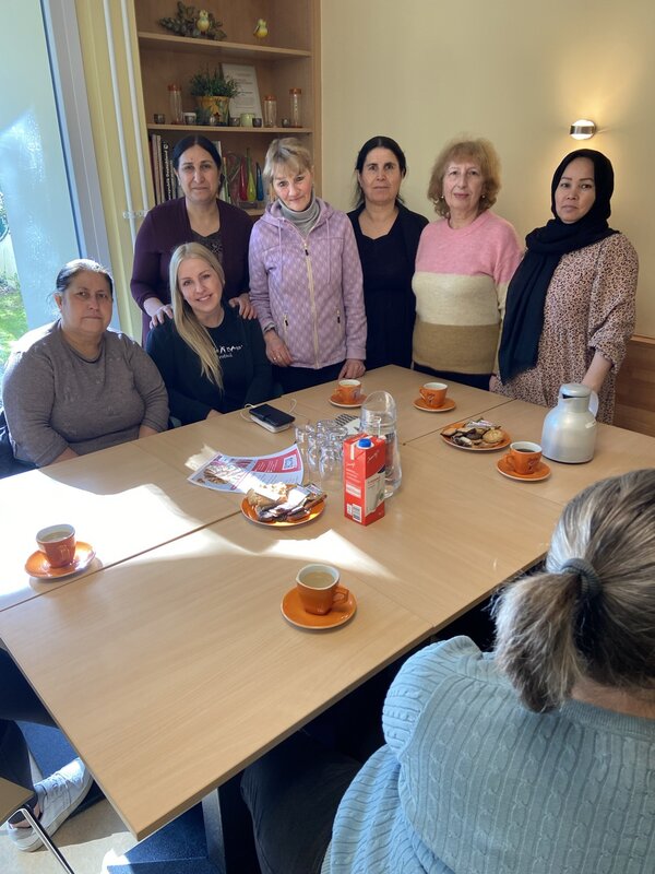 Eine Gruppe von acht Frauen sitzt und steht um einen Tisch, auf dem Kaffee, Gebäck und ein Flyer liegen. Sie blicken in die Kamera, einige von ihnen lächeln. Im Hintergrund befinden sich Regale mit Dekorationsgegenständen.