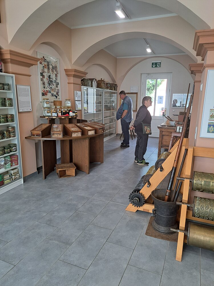 Blick in einen Museumsraum mit historischen Ausstellungsstücken wie Dosen, Werkzeugen und Verpackungen. Zwei Besucher betrachten die Exponate an den Vitrinen.