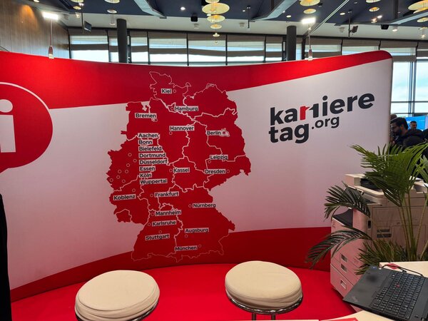 Roter Messestand „karrieretag.org“ mit Deutschlandkarte und markierten Städten; davor zwei Hocker, Pflanzen und Technik.