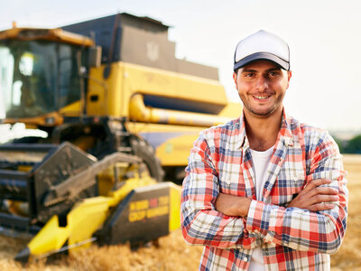 Ein lächelnder Mann mit einer Basecap steht verschränkt vor einem gelben Mähdrescher auf einem Getreidefeld. Er trägt ein kariertes Hemd und wirkt selbstbewusst. Der Mähdrescher ist im Hintergrund leicht unscharf zu sehen, während das Feld golden schimmert.