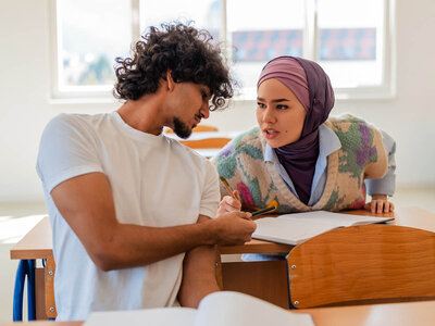 Ein Mann mit lockigem Haar und einem weißen T-Shirt sitzt an einem Schreibtisch und schreibt in ein Notizbuch. Neben ihm steht eine Frau mit einem lila Kopftuch und einem gemusterten Cardigan. Sie beobachtet, was er schreibt. Beide wirken konzentriert. Im Hintergrund sind weitere Schreibtische und Fenster zu sehen.