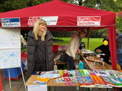 Zwei Mitarbeiterinnen stehen am Euro-Schulen Stand, der mit Kuchen sowie Lehrmaterialien gefüllt ist. Zwei Besucher holen sich ein Stück Kuchen.