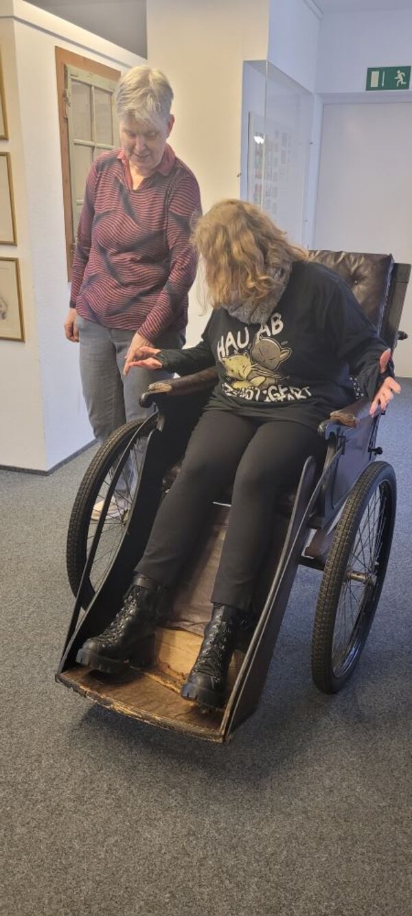 Zwei Besucherinnen erkunden einen historischen Rollstuhl. Eine davon sitzt in dem Rollstuhl, während die andere auf eins der Räder deutet.