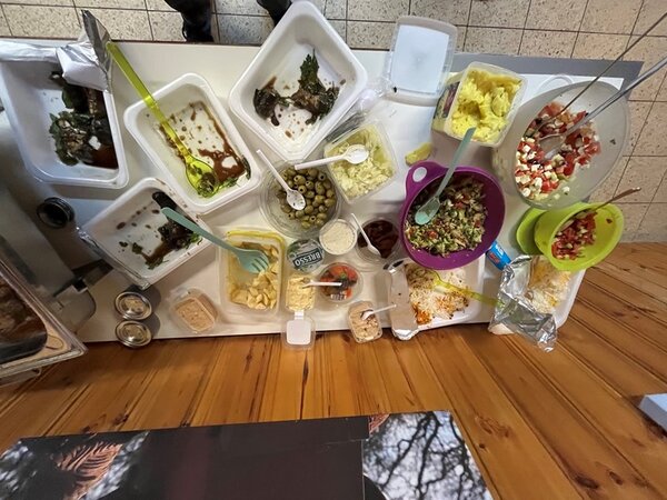 Verschiedene Schalen und Behälter mit Salaten, Oliven, Käse, Saucen und weiteren Lebensmitteln stehen auf einem Tisch. Einige Schalen sind aus Kunststoff, andere aus Metall. Besteck wie Löffel und Zangen liegen in den Behältern. Die Oberfläche des Tisches ist teilweise aus Holz, daneben sind Fliesen zu sehen. Die Szene zeigt eine Auswahl an Speisen, die zum Selbstbedienen bereitgestellt sind.