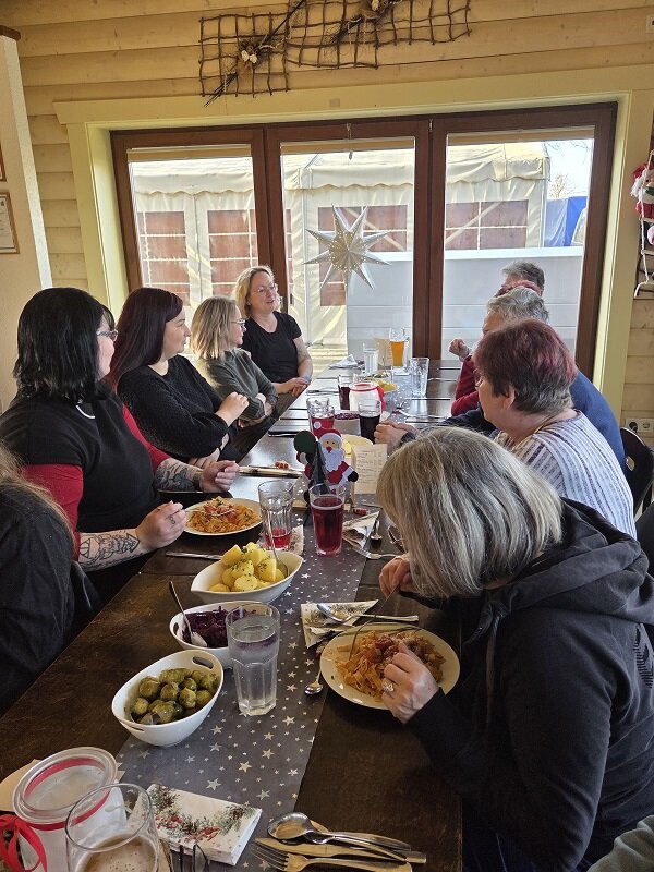 Mehrere Personen sitzen dicht an einem Tisch und essen gemeinsam, auf dem Tisch stehen Getränke, Teller mit warmen Speisen und weihnachtliche Dekoration