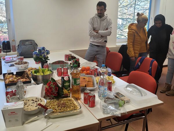 In einem Unterrichtsraum steht ein langer Tisch voll mit selbst mitgebrachten Speisen, Getränken und Blumen; mehrere Kursteilnehmende stehen daneben und betrachten das Buffet.