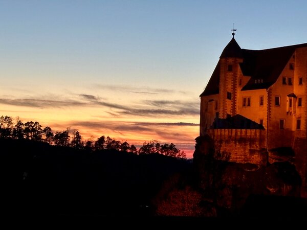 Historisches Schloss auf einem Felsen bei Sonnenuntergang, goldene Lichtstimmung am Himmel mit silhouettiertem Wald im Hintergrund.