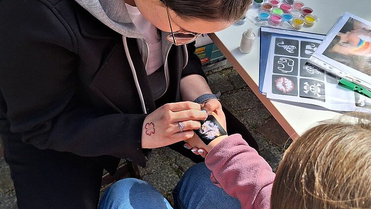 Mit Herz und Kreativität dabei Eine Frau klebt ein Tattoo auf die Hand eines Kindes. Auf dem Tisch liegen Schablonen und Farben für das Schminken.