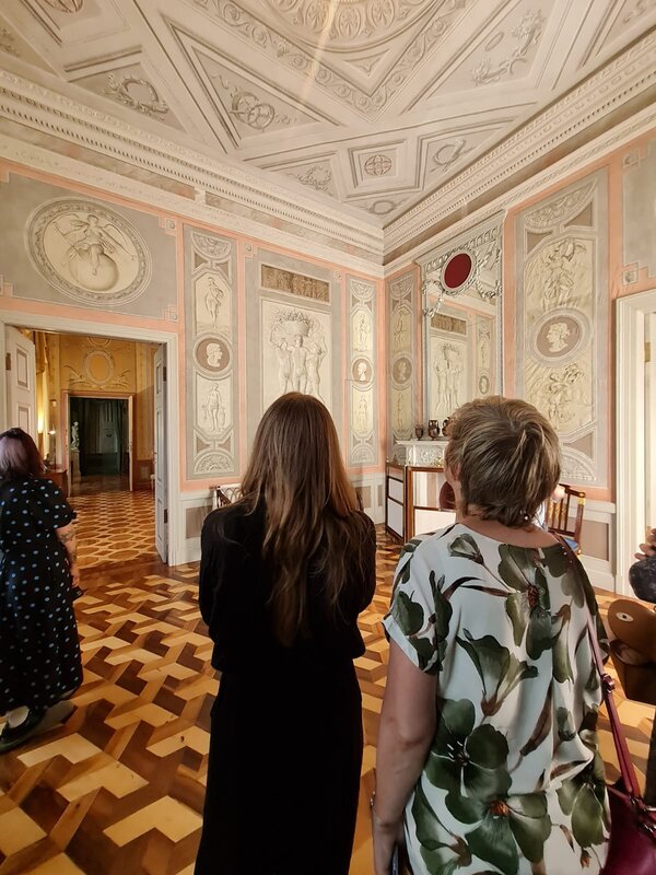 Zwei Personen sind von hinten in einem auffällig verzierten Raum in einem Schloss zu sehen. Sie schauen beide nach oben und betrachten die verzierte Decke.