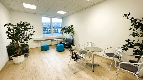 Gemütlicher Ruheraum mit zwei großen Zimmerpflanzen, kleinen runden Metalltischen, Stühlen, zwei Sitzbänken und türkisfarbenen Sitzsäcken vor einem Fenster.