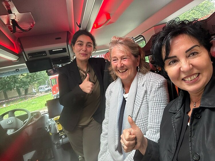 Drei lachende Frauen in einem Einsatzfahrzeug der Feuerwehr zeigen "Daumen hoch" in die Kamera.
