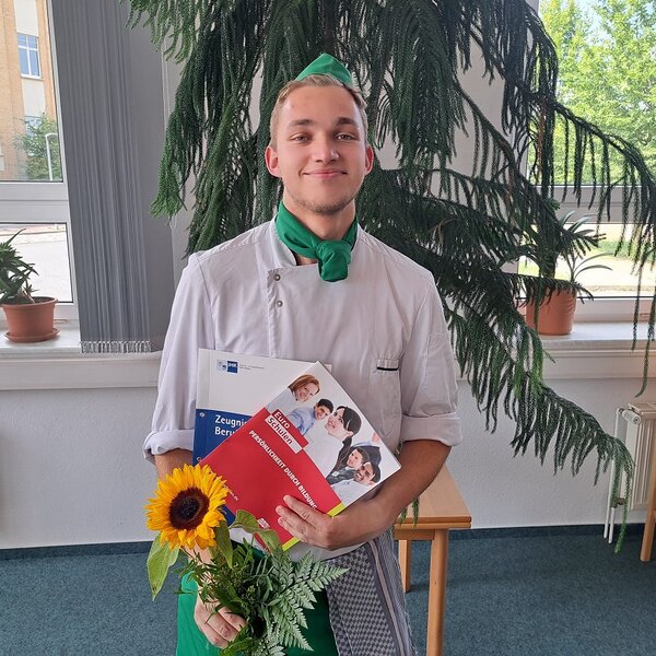 Ein Absolvent grinst zufrieden in die Kamera. Er trägt ein Kochoutfit mit grüner Schürze, Halstuch und Kochmütze. In den Händen hält er die Prüfungszeugnisse in Mappen der IHK und der Euro-Schulen sowie einen Strauß mit einer Sonnenblume.