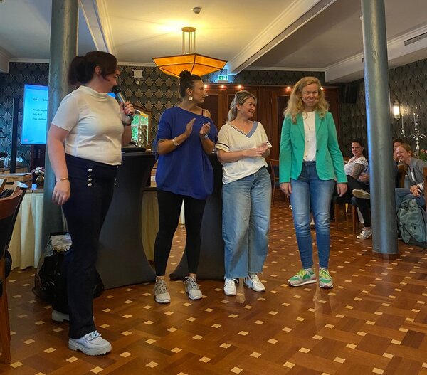 Vier Frauen stehen in einem Raum mit dekorativen Wandmustern und einer auffälligen Designerleuchte. Eine Frau hält ein Mikrofon, während die anderen drei Frauen lächeln und eine klatscht. Im Hintergrund sind weitere Personen, die sitzen zu sehen.