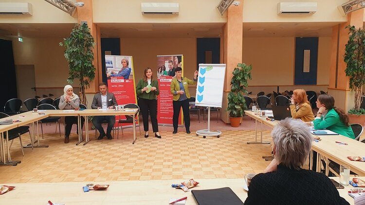 Teilnehmende sitzen an Tischen und schauen interessiert nach vorn zu den zwei weiblichen Teilnehmenden, die an einem Flipchart Ergebnisse präsentieren.