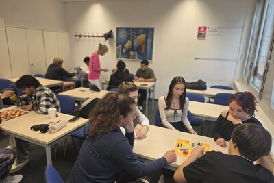 Mehrere Gruppen von Jugendlichen sitzen in einem Klassenraum und spielen verschiedene Gesellschaftsspiele, darunter „Mensch ärgere dich nicht“ und Dame. Eine Lehrkraft steht im Hintergrund und beobachtet.