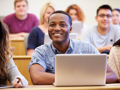 Ein lächelnder Mann mit einem hellblauen Hemd sitzt in einem Hörsaal und arbeitet an einem Laptop. Im Hintergrund sind mehrere Personen zu sehen, die ebenfalls an Tischen sitzen und arbeiten oder zuhören.