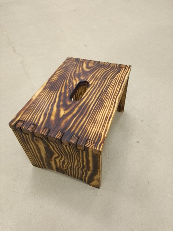 Kleine geflammte Holz-Fußbank mit Tragegriff-Ausschnitt in der Sitzfläche; dunkle Maserung und sichtbare Holzverbindungen auf hellem Boden.