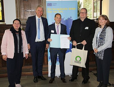 Euro Akademie Oldenburg: weiterhin „die“ Europaschule in Niedersachsen Mehrere Politiker*innen und der Schulleiter Ludwig Knapp haben sich für ein Foto positioniert, dabei wir die Urkunde "Europaschule in Niedersachsen" übergeben. Die Szene spielt sich im Osnabrücker Rathaus ab, hinter den Personen steht ein Rollup, das für die Europäische Union wirbt.
