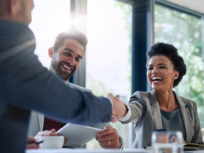 Drei Personen sitzen an einem Tisch und eine schwarzhaarige Frau gibt einem Mann die Hand.