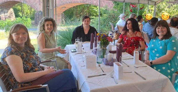 Eine Gruppe von sechs Personen sitzt an einem langen, festlich gedeckten Tisch auf einer Terrasse mit Blick auf einen grünen Garten und historische Backsteinbögen. Auf dem Tisch stehen Blumen, Gläser und Geschenktüten. Die Personen lächeln in die Kamera.