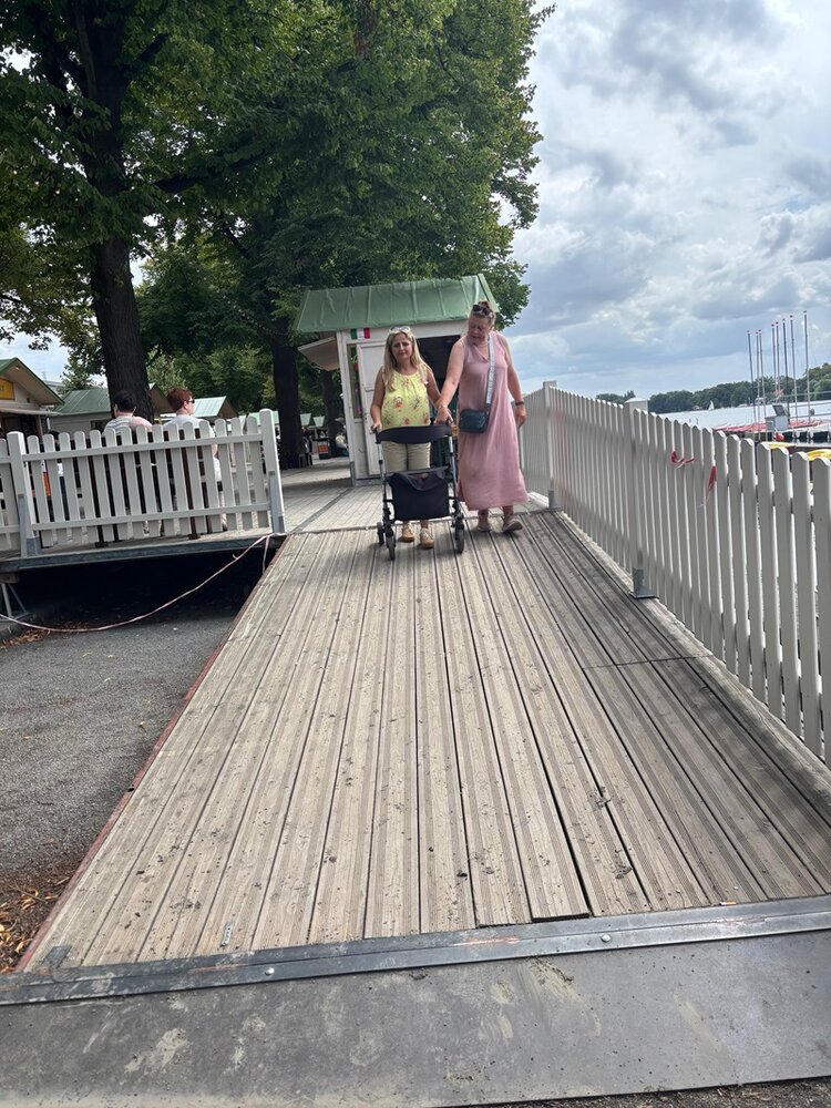 Im Vordergrund verläuft eine breite Rampe aus hellem Holz, die von einem weißen Geländer eingefasst wird. Zwei Frauen gehen nebeneinander die Rampe hinauf, eine schiebt einen Kinderwagen. Im Hintergrund stehen Bäume und ein kleiner Pavillon mit grünem Dach. Rechts sind Segelboote am Ufer zu erkennen.