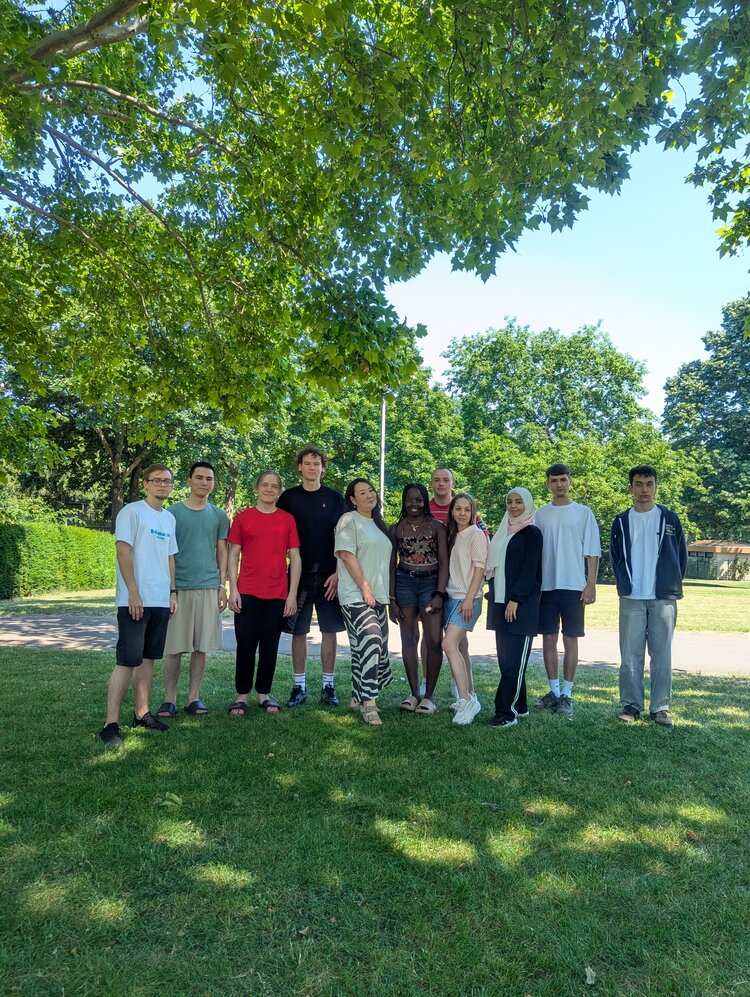 Eine Gruppe von elf jungen Erwachsenen steht gemeinsam auf einer Wiese unter großen, grünen Bäumen in einem sonnigen Park.