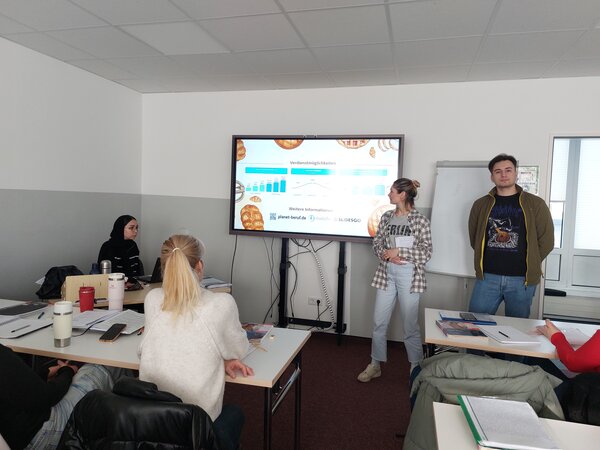 Eine junge Frau und ein junger Mann präsentieren vor einer Klasse Informationen zu Verdienstmöglichkeiten. Der Bildschirm zeigt ein Diagramm sowie Logos für weitere Informationsquellen. Die Teilnehmenden hören aufmerksam zu.