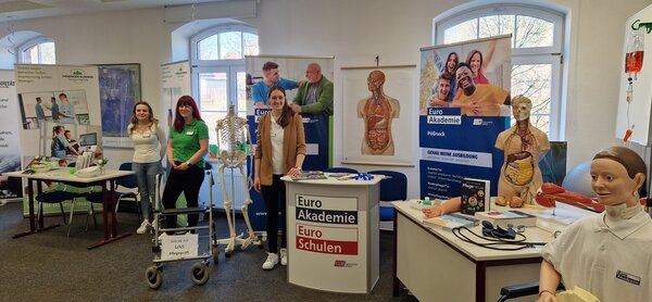 Ein Blick auf die gleiche Ausstellungsfläche aus einer leicht anderen Perspektive. Im Vordergrund ein Anatomie-Modell und ein Rollator mit dem Schild „Werde mit UNS Pflegeprofi“. Freundliche Standbetreuerinnen stehen bereit.