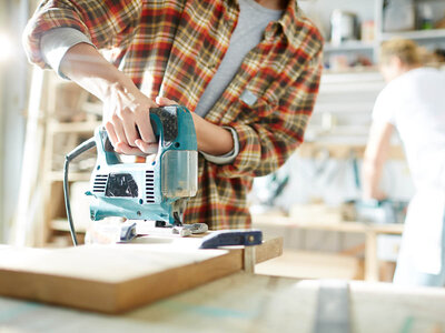 Eine Person mit einem karierten Hemd arbeitet mit einer elektrischen Stichsäge an einem Holzbrett in einer Werkstatt oder einem Arbeitsraum. Die Hände der Person sind auf der Säge platziert, um einen präzisen Schnitt durchzuführen. Im Hintergrund ist eine weitere Person zu sehen, die sich in der Nähe von Regalen und Werkzeugen befindet. Der Raum ist gut beleuchtet und scheint auf handwerkliche Arbeiten spezialisiert zu sein.